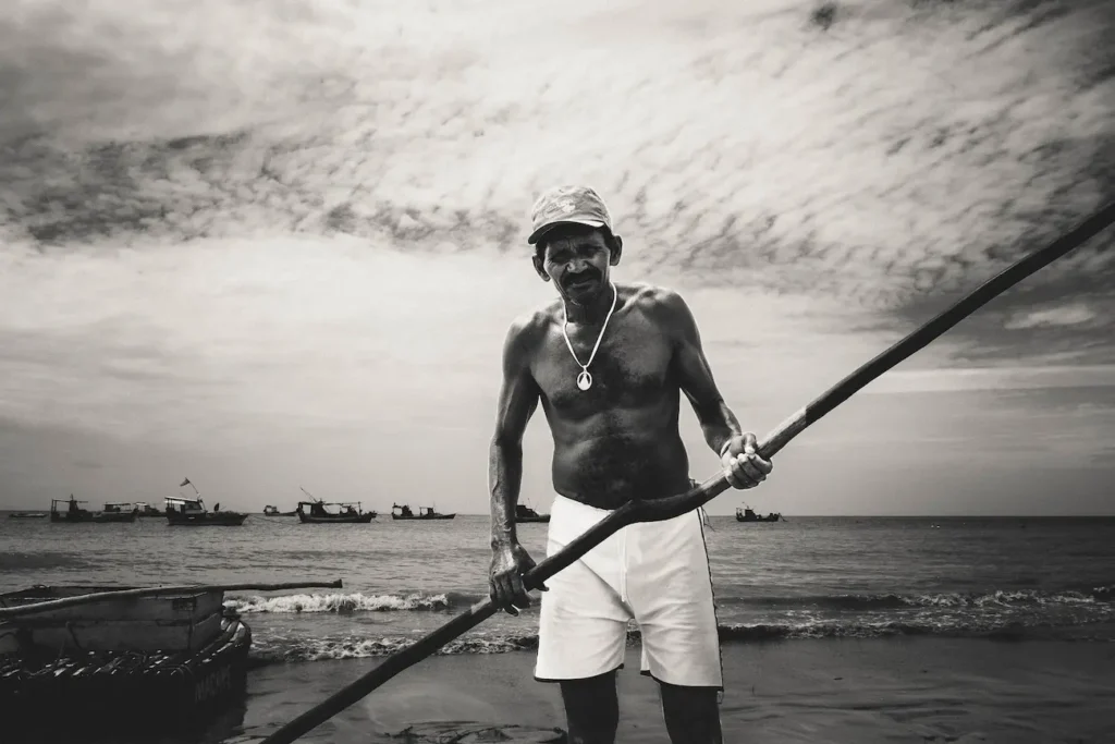 Costa del Nordeste, Brasile: ritratto in bianco e nero di un pescatore anziano a torso nudo sulla spiaggia. Indossa un cappello e tiene un remo, con barche da pesca sullo sfondo. Foto di Filippo Cristallo, dal progetto "Chuva Suor e Cherveza
