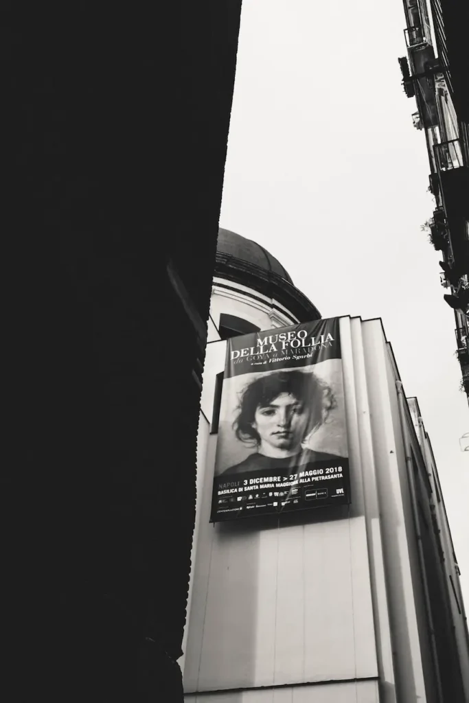 Manifesto della mostra “Museo della Follia” appeso alla facciata di un edificio tra due palazzi nel centro storico di Napoli.