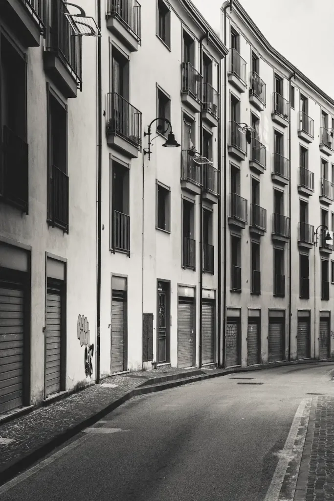 Strada curva tra edifici residenziali, balconi ripetuti e serrande abbassate.
