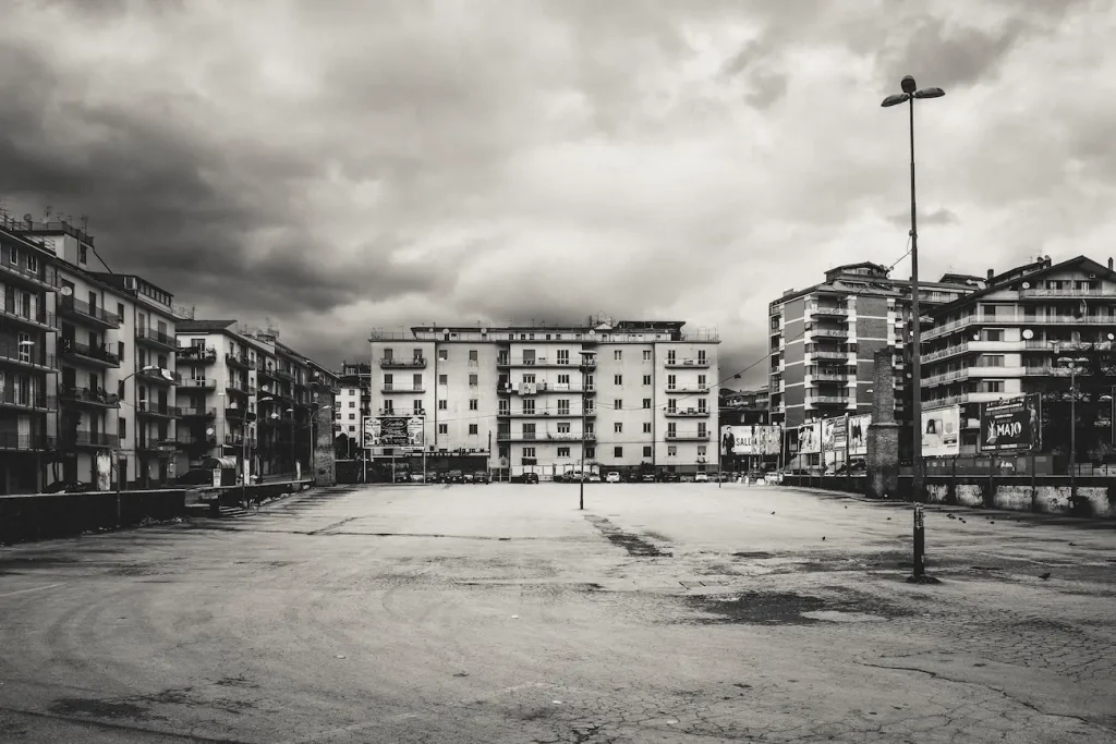 Spiazzo deserto, palazzi bassi e cartelloni segnano un margine urbano sospeso.