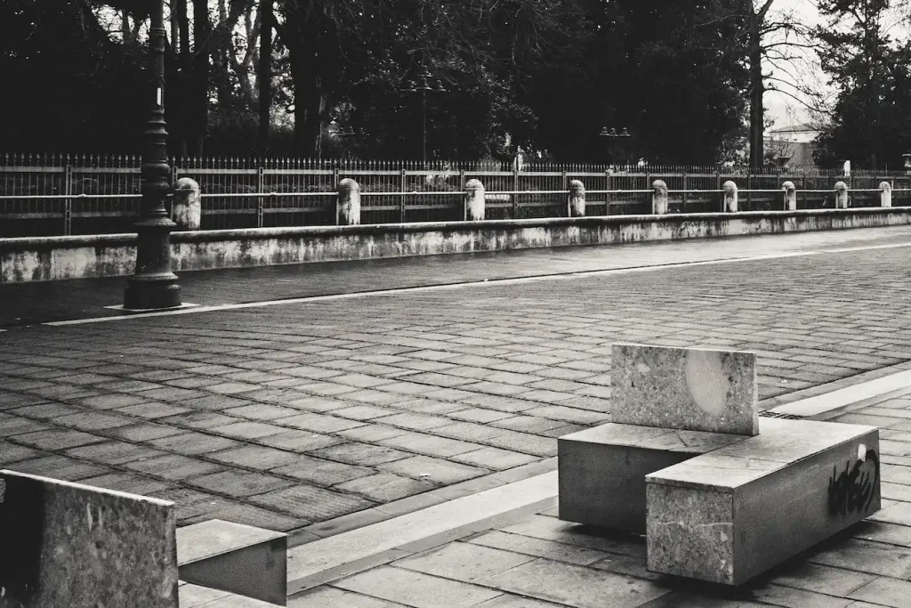 Sedute in pietra e pavimentazione deserta davanti a una cancellata urbana silenziosa.